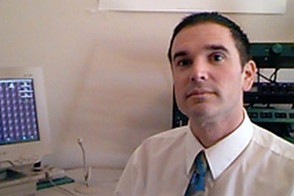Michael McAnally at his desk in a shirt and tie, a computer monitor beside him